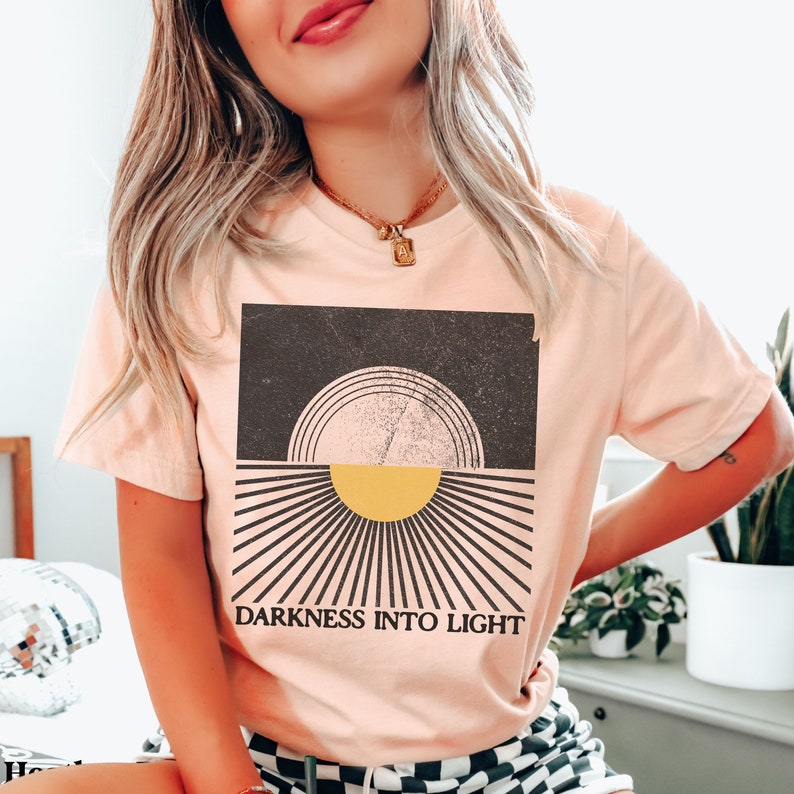May include: A pink t-shirt with a graphic design of a sun rising over a dark background. The graphic has a vintage, distressed look. The text "DARKNESS INTO LIGHT" is printed below the graphic.