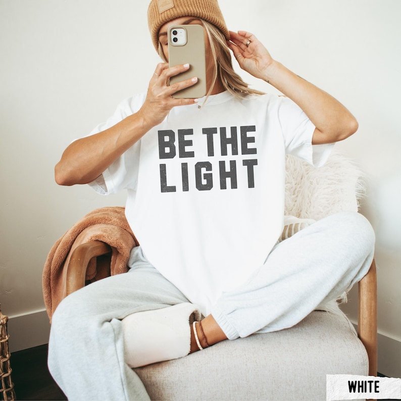Peut inclure: T-shirt blanc avec le texte "BE THE LIGHT" en gras, en gris fonc&eacute;. La personne porte un bonnet beige, un pantalon de surv&ecirc;tement gris et des chaussettes blanches. La personne est assise sur une chaise et tient un t&eacute;l&eacute;phone.