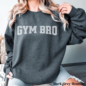 May include: Dark grey heather sweatshirt with the text "GYM BRO" in a light grey, block letter font. The sweatshirt is a crew neck style and appears to be made of a soft material. The words "Dark Grey Heather" are printed at the bottom.