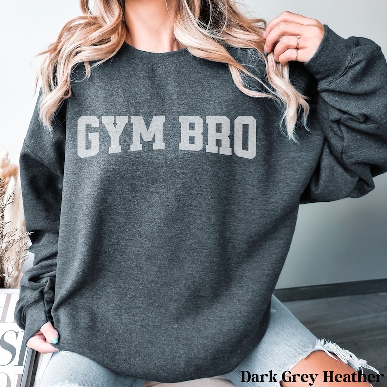May include: Dark grey heather sweatshirt with the text "GYM BRO" in a light grey, block letter font. The sweatshirt is a crew neck style and appears to be made of a soft material. The words "Dark Grey Heather" are printed at the bottom.