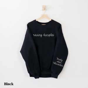 May include: Black sweatshirt with the words "raising disciples" in white script across the chest. The sleeve features the names "brody eden alexander" in white script. The word "Black" is at the bottom.