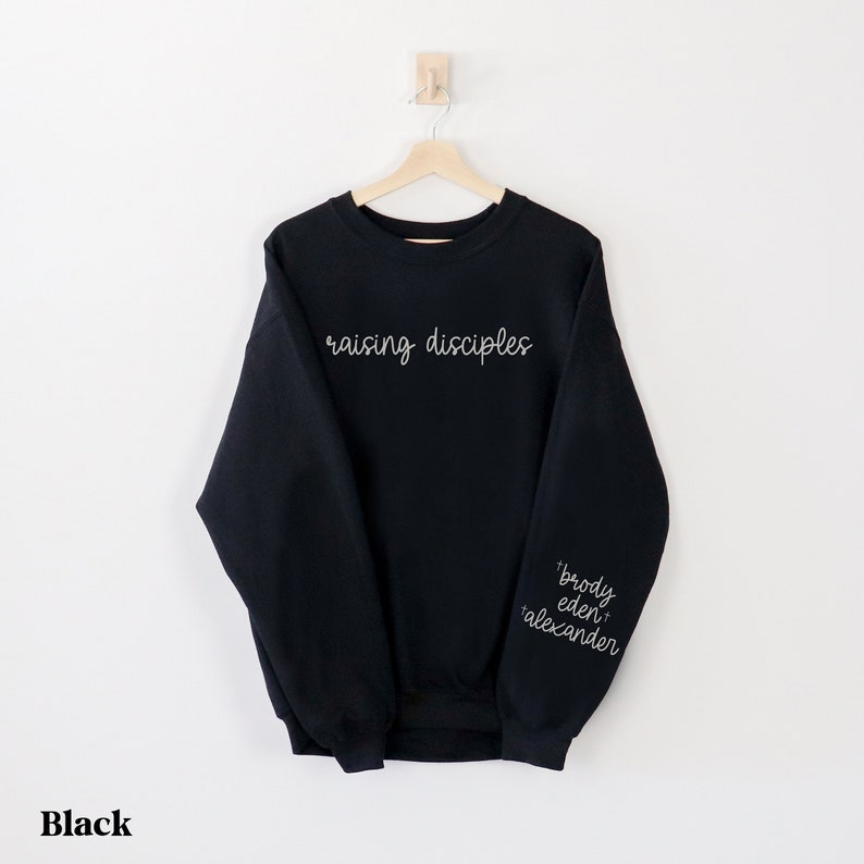 May include: Black sweatshirt with the words "raising disciples" in white script across the chest. The sleeve features the names "brody eden alexander" in white script. The word "Black" is at the bottom.