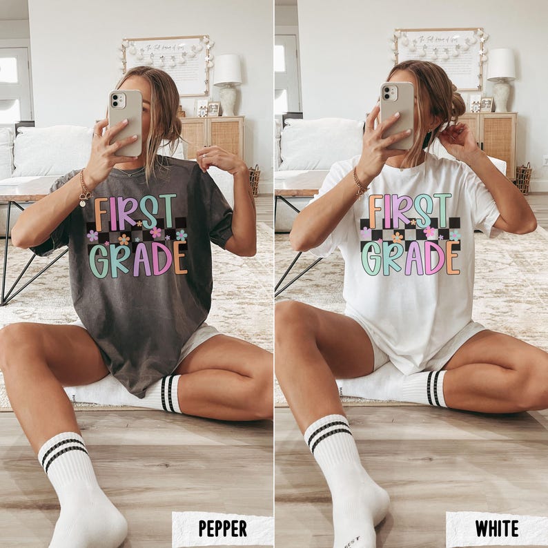 K&ouml;nnte beinhalten: Zwei T-Shirts, eines in Grau und eines in Wei&szlig;, mit den Worten "FIRST GRADE" in bunten Buchstaben und einem Schachbrettmuster. Die Shirts sind kurz&auml;rmlig und werden mit wei&szlig;en Shorts und Socken getragen.