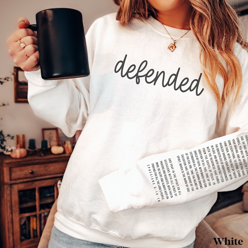 May include: White sweatshirt with the word "defended" in a script font. The right sleeve features a printed passage from Ephesians 6:10-18. A black mug is held in the left hand.
