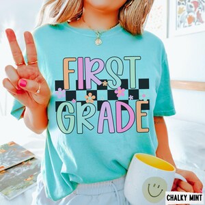 K&ouml;nnte beinhalten: Ein hellt&uuml;rkisfarbenes T-Shirt mit den Worten "FIRST GRADE" in bunten, umrandeten Buchstaben. Der Text befindet sich &uuml;ber einem schwarz-wei&szlig;en Schachbrettmuster mit Blumenakzenten. Eine wei&szlig;e Tasse mit gelbem Innenraum und Smiley ist ebenfalls zu sehen.