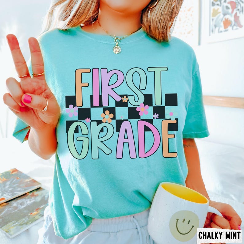 K&ouml;nnte beinhalten: Ein hellt&uuml;rkisfarbenes T-Shirt mit den Worten "FIRST GRADE" in bunten, umrandeten Buchstaben. Der Text befindet sich &uuml;ber einem schwarz-wei&szlig;en Schachbrettmuster mit Blumenakzenten. Eine wei&szlig;e Tasse mit gelbem Innenraum und Smiley ist ebenfalls zu sehen.