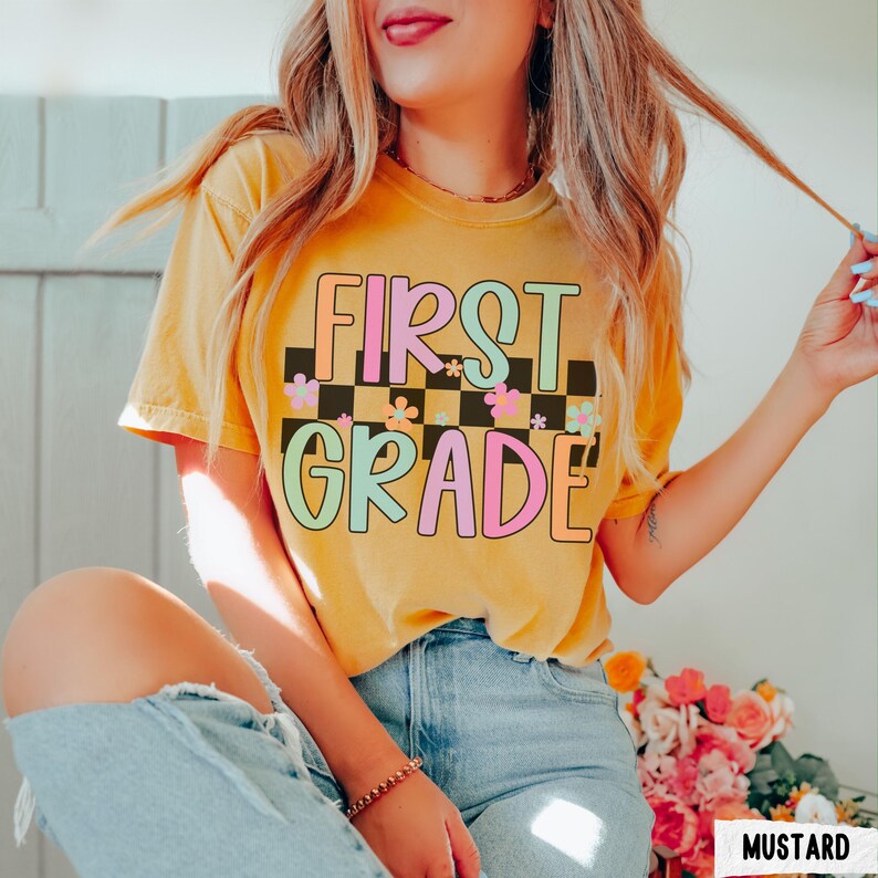 K&ouml;nnte beinhalten: Senffarbenes T-Shirt mit dem Schriftzug "FIRST GRADE" in einer farbenfrohen Retro-Schriftart. Der Text ist vor einem schwarz-wei&szlig; karierten Hintergrund mit floralen Akzenten platziert. Das Shirt ist kurz&auml;rmelig.