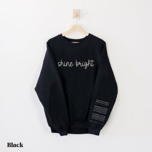 May include: Black sweatshirt with the text "shine bright" printed on the front. The sleeve has a quote from Matthew 5:16.