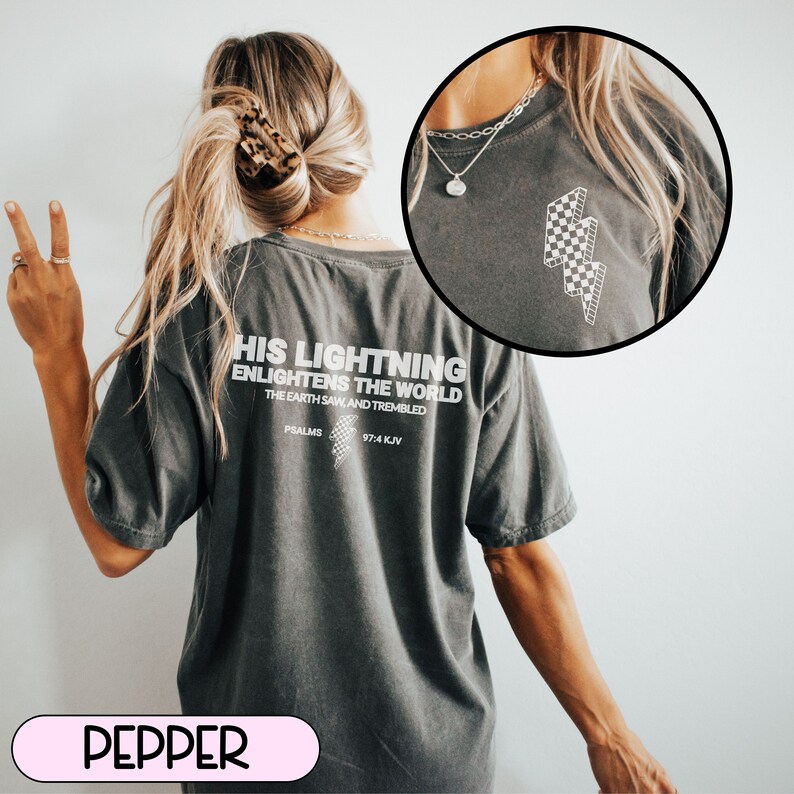 May include: A dark gray t-shirt with white text on the back that reads "HIS LIGHTNING ENLIGHTENS THE WORLD." The shirt also features a checkered lightning bolt design. The text "PSALMS 97:4 KJV" is below the main text.