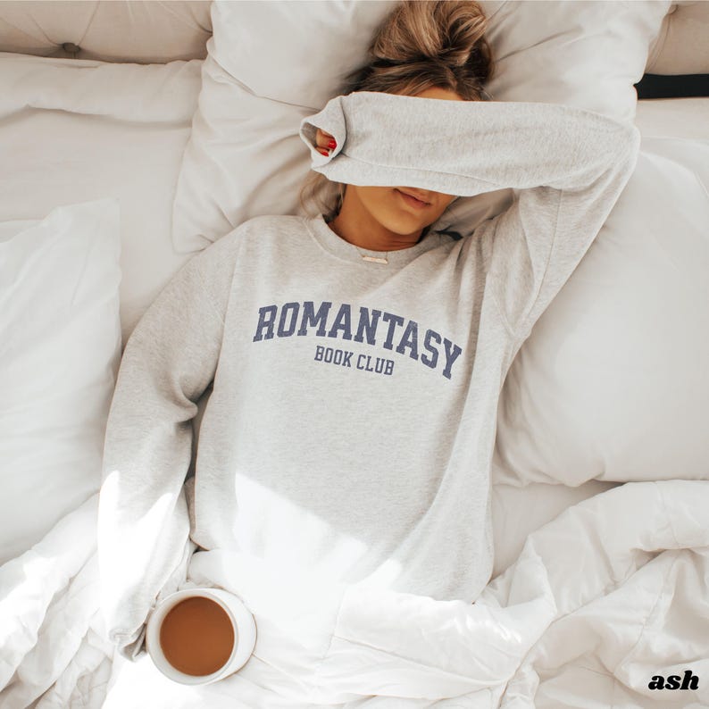 May include: A grey sweatshirt with the text "ROMANTASY BOOK CLUB" in blue lettering. The sweatshirt is being worn by a person lying in bed.