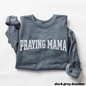 Pode incluir: Uma camisa de moletom cinza escuro com o texto "Praying Mama" impresso em branco.