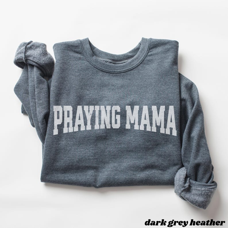 Pode incluir: Uma camisa de moletom cinza escuro com o texto "Praying Mama" impresso em branco.