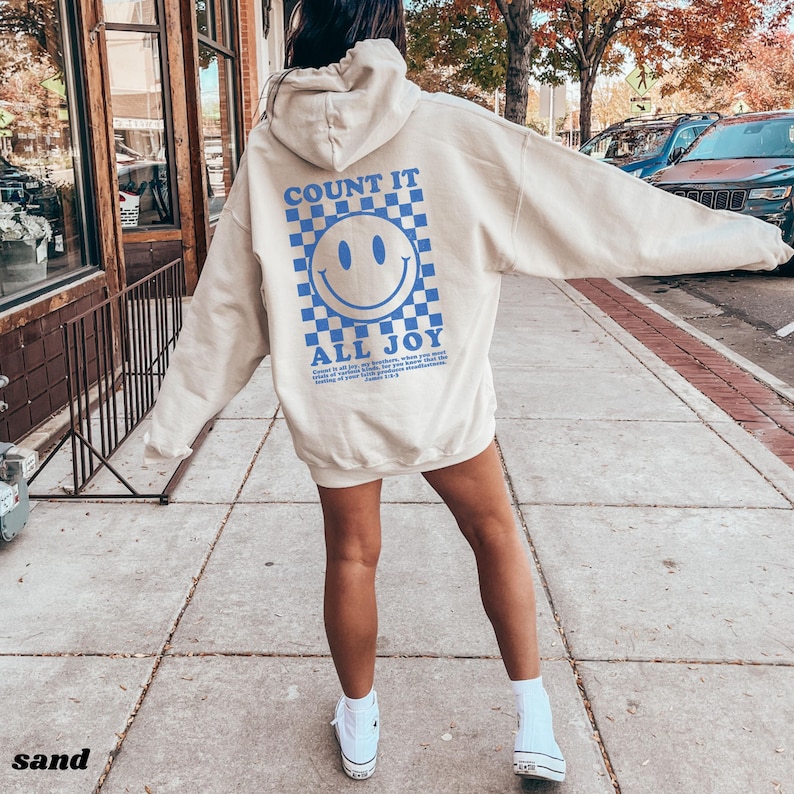 May include: A beige hoodie with a blue checkered pattern and a blue smiley face graphic. The text "COUNT IT ALL JOY" is printed below the graphic. The hoodie is being worn by a person.