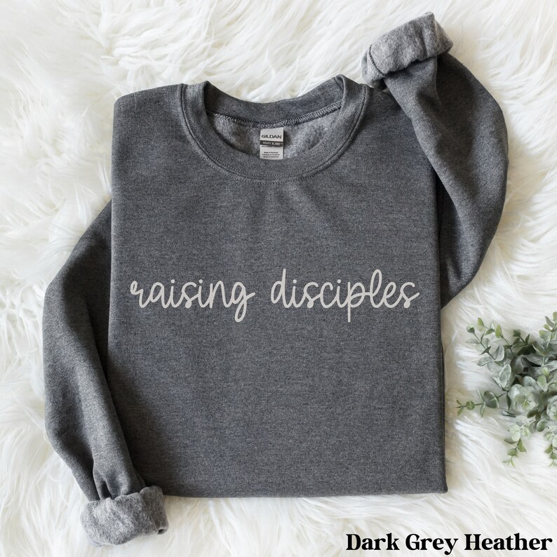 May include: Dark grey heather sweatshirt with the phrase "raising disciples" in a cursive font. The long-sleeved top is made of a soft material and has a crew neck. The sleeves are cuffed at the wrists.