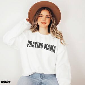 Pode incluir: Camisola branca com o texto "Praying Mama" em letras pretas. A modelo est&aacute; usando um chap&eacute;u castanho e cal&ccedil;as de ganga azuis.