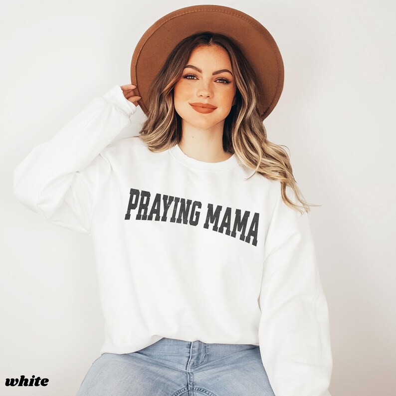 Pode incluir: Camisola branca com o texto "Praying Mama" em letras pretas. A modelo est&aacute; usando um chap&eacute;u castanho e cal&ccedil;as de ganga azuis.