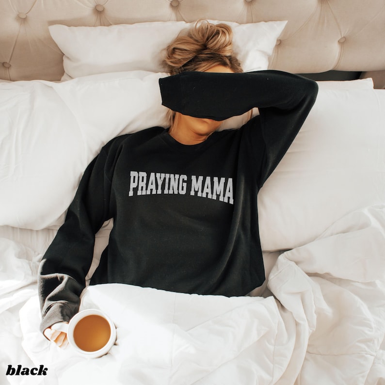 Pode incluir: Uma camisola preta com o texto "Praying Mama" em letras brancas. A camisola &eacute; usada por uma pessoa deitada na cama.