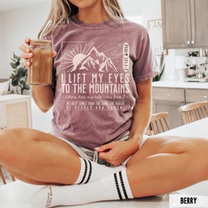 May include: Berry-colored t-shirt with the text "I Lift My Eyes to the Mountains" and a mountain graphic. The shirt also includes the text "Psalm 121:1-2" and "My help comes from the Lord, the maker of heaven and earth." The person is holding a glass of iced coffee.