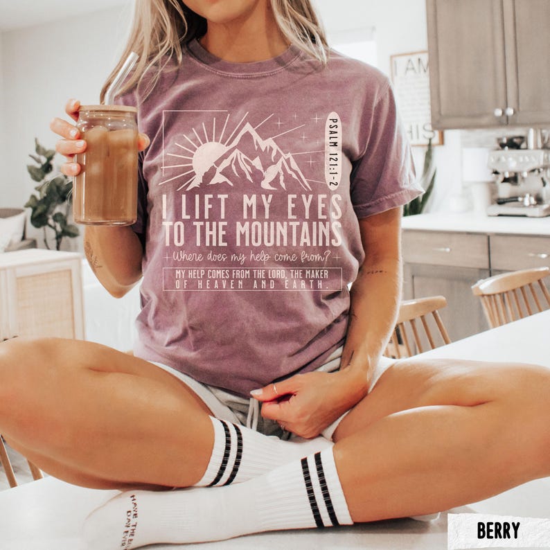 May include: Berry-colored t-shirt with the text "I Lift My Eyes to the Mountains" and a mountain graphic. The shirt also includes the text "Psalm 121:1-2" and "My help comes from the Lord, the maker of heaven and earth." The person is holding a glass of iced coffee.