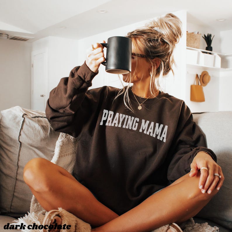 Pode incluir: Uma camisa de moletom marrom com o texto "Praying Mama" em letras brancas. A modelo est&aacute; vestindo a camisa de moletom e segurando uma caneca preta.