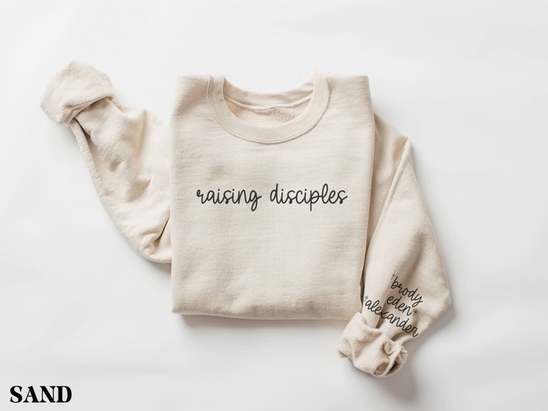 May include: Sand-colored sweatshirt with the words "raising disciples" in black script across the chest. The sleeve cuff features the names "brody eden alexander" in black script. The sweatshirt is folded.