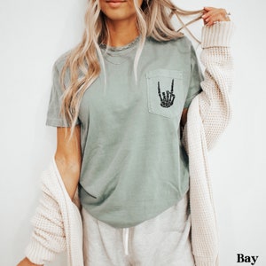 May include: A sage green t-shirt with a black skeleton hand making a rock and roll sign in a pocket on the front of the shirt.