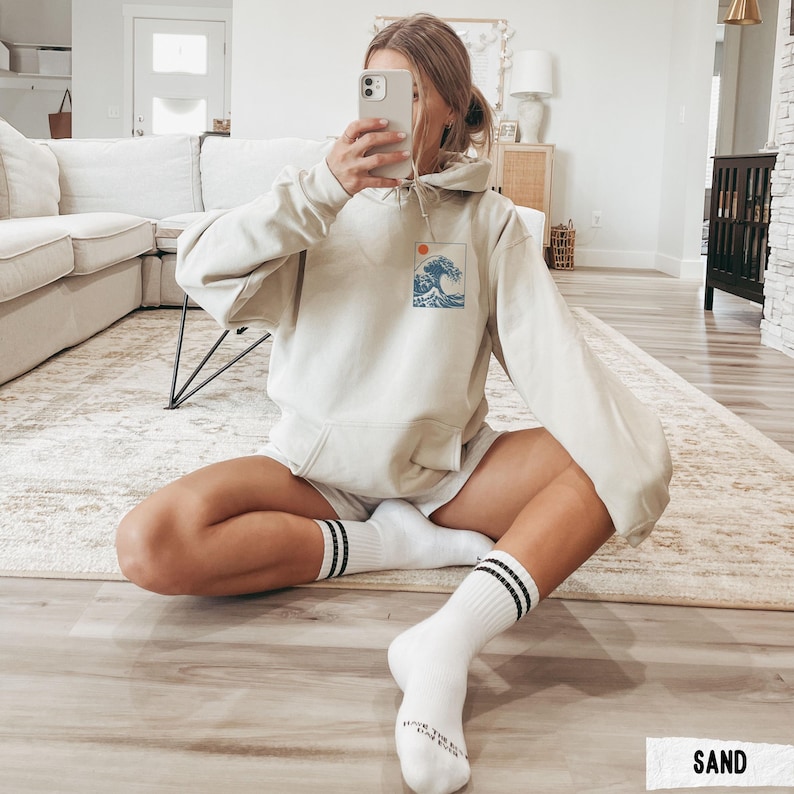 May include: A sand-colored hoodie with a front pocket and a drawstring hood. The hoodie features a small graphic of a wave and sun. The person is wearing white shorts and white socks with black stripes.
