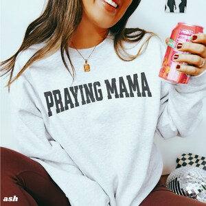 Pode incluir: Uma camisa de moletom branca com o texto "Praying Mama" impresso em preto na frente. A camisa de moletom est&aacute; sendo usada por uma pessoa.