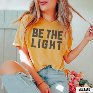 Peut inclure: T-shirt jaune moutarde avec l'inscription "BE THE LIGHT" en lettres majuscules gris fonc&eacute;. Le t-shirt est &agrave; manches courtes et a un col rond. Le mod&egrave;le porte un jean bleu clair. Le mot "MUSTARD" est dans un encadr&eacute; blanc.