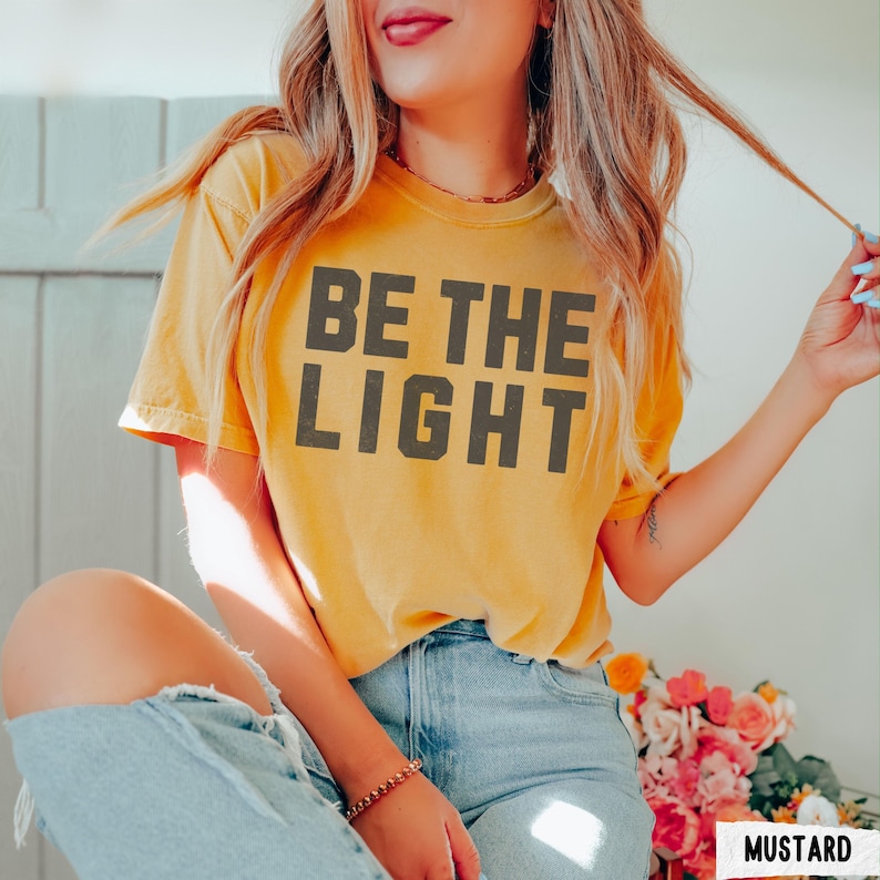 Peut inclure: T-shirt jaune moutarde avec l'inscription "BE THE LIGHT" en lettres majuscules gris fonc&eacute;. Le t-shirt est &agrave; manches courtes et a un col rond. Le mod&egrave;le porte un jean bleu clair. Le mot "MUSTARD" est dans un encadr&eacute; blanc.
