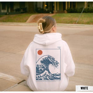 May include: White hoodie featuring a blue wave graphic with a red sun and text. The design is framed in a navy blue rectangle. The hoodie is a casual garment, suitable for everyday wear.