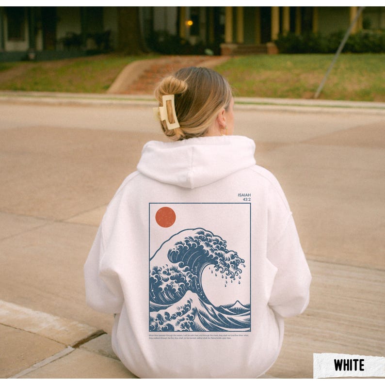 May include: White hoodie featuring a blue wave graphic with a red sun and text. The design is framed in a navy blue rectangle. The hoodie is a casual garment, suitable for everyday wear.