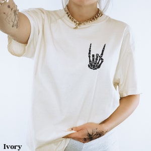 May include: Ivory t-shirt with a black skeleton hand making a rock and roll sign.
