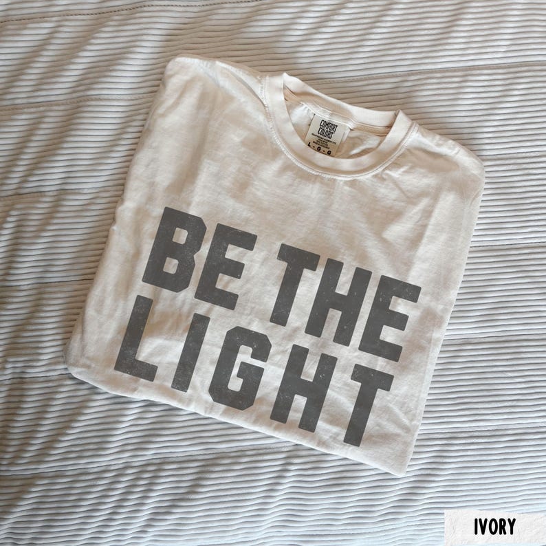 May include: Ivory-colored t-shirt with the phrase "BE THE LIGHT" printed in gray block letters. The shirt is folded and resting on a striped, light-colored fabric. The tag reads "Comfort Colors".