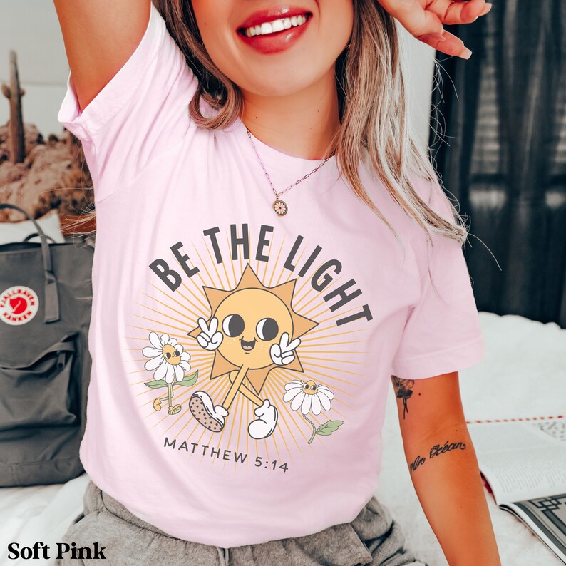 Op de afbeelding: Een zacht roze t-shirt met een cartoon zon-afbeelding en de tekst "Wees het licht" met de Bijbeltekst "Matthe&uuml;s 5:14".