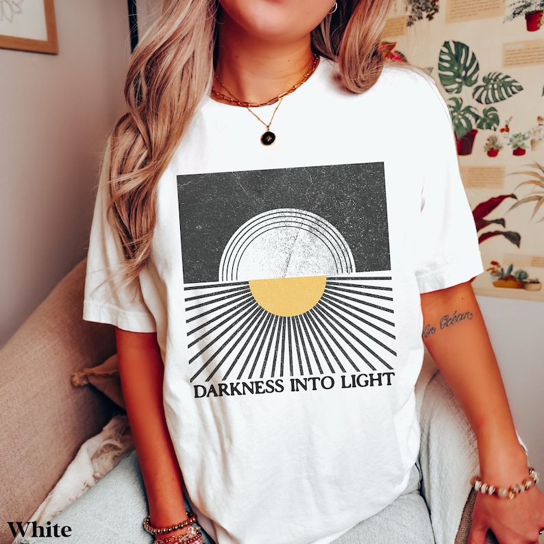 May include: White t-shirt with a graphic design of a sun rising over a black and white striped background. The text "DARKNESS INTO LIGHT" is printed below the graphic.