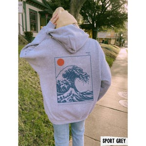 May include: Sport grey hoodie with a large graphic on the back featuring a blue wave design, a red sun, and text. The hoodie has a hood and is worn with blue jeans. The text "SPORT GREY" is at the bottom of the image.