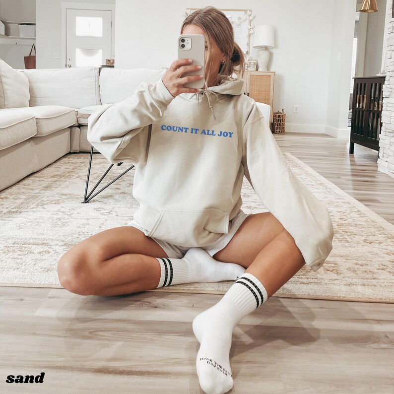 May include: Sand-colored hoodie with the text "COUNT IT ALL JOY" in blue. The person is wearing white shorts and white socks with black stripes. The word "sand" is at the bottom left of the image.