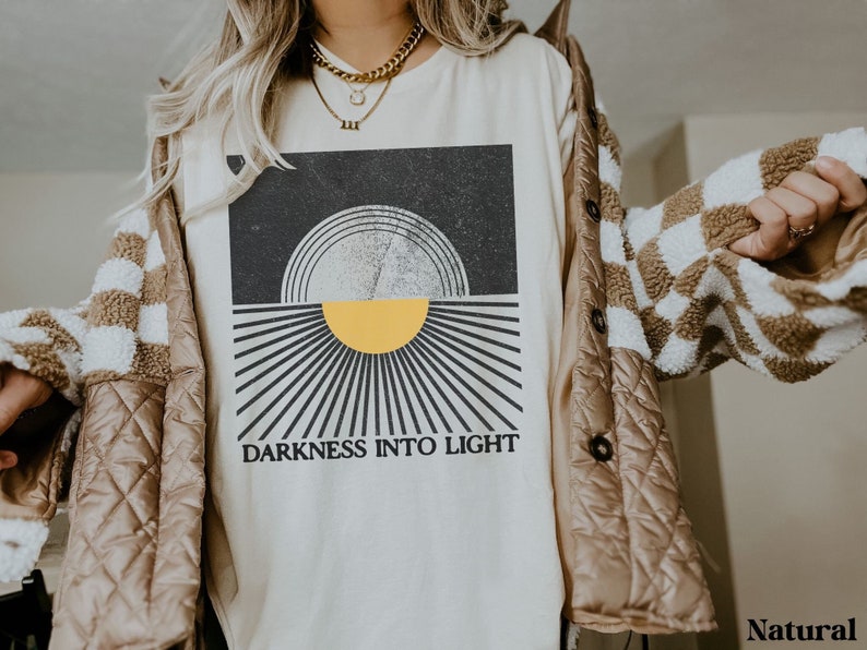 May include: A white t-shirt with a black and white graphic of a sun with the text "Darkness into Light".