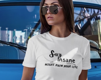 May include: White t-shirt with the text "Sexy Insane Might Ruin Your Life" in black script. The shirt has a crew neck and short sleeves. The model is wearing sunglasses.