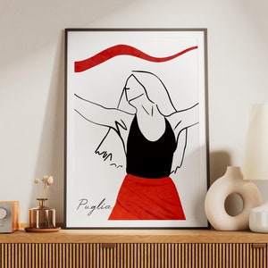 Può includere: Una stampa artistica incorniciata con un'illustrazione minimalista di una donna con top nero e gonna rossa, con la parola "Puglia" sotto. L'opera d'arte è esposta su un mobile in legno con stereo, vaso, lampada e oggetti decorativi.