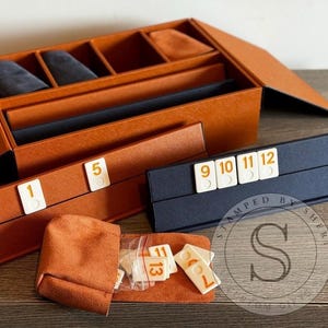May include: A brown leather box with compartments for storing game pieces. The box is open and the compartments are filled with small, white tiles with numbers on them. The tiles are arranged in a row, with the numbers 1, 5, 9, 10, 11, and 12 visible. The box is also labeled "Stamped by Sherry".