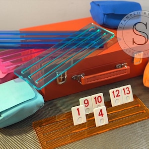 Op de afbeelding: Een oranje acryl Rummikub-tegelhouder met witte tegels met de nummers 1, 4, 9, 10, 12 en 13. De afbeelding toont ook kleurrijke acrylhouders en kleine tassen in blauw, roze en oranje.