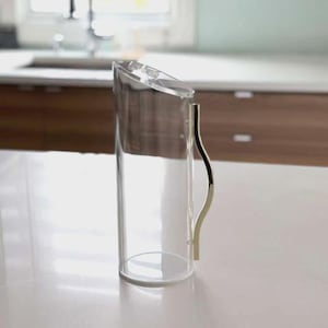 May include: A clear, cylindrical pitcher with a slanted top and a gold-colored handle. The pitcher is made of glass and sits on a white surface. The handle has a curved design.