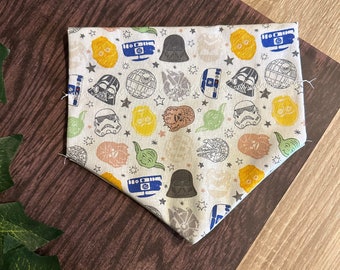 May include: A beige fabric bandana with a white background and a repeating pattern of Star Wars characters, including Darth Vader, Yoda, and R2-D2. The bandana has a blue, yellow, and white color scheme.