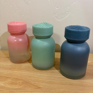 Puede incluir: Tres pequeñas botellas cilíndricas en rosa, verde y azul. Cada botella tiene una parte superior redondeada y una tapa a juego con un diseño texturizado. Las botellas se muestran sobre una superficie de madera marrón claro. Las botellas miden unos 10 cm de alto.