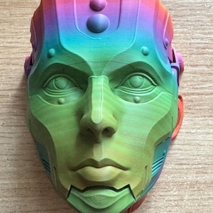 May include: A 3D printed mask with a rainbow gradient on the top and a green face with detailed features. The mask has a futuristic design with a textured surface.