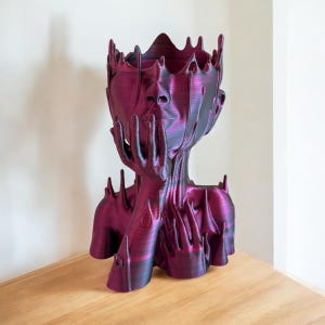 May include: A 3D printed sculpture of a human head with hands, made in a deep purple colour with a metallic sheen. The head is tilted down and the hands are clasped together in front of the face. The sculpture is a unique and modern piece of art.