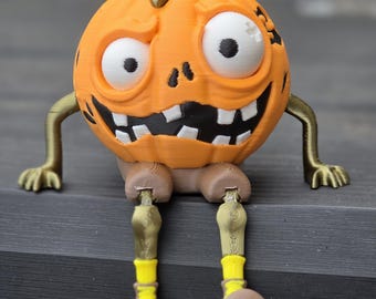 Puede incluir: Una figura de calabaza naranja impresa en 3D con una cara de dibujos animados, dientes blancos y ojos grandes blancos. Tiene piernas y brazos articulados en tonos de marrón, dorado y amarillo. La calabaza es una decoración de Halloween.