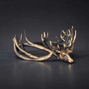 May include: A circular bracelet featuring a sculpted deer head with prominent antlers. The accessory is finished in a metallic gold and black colour scheme, highlighting the textured details of the animal's features and the branching antler structure.